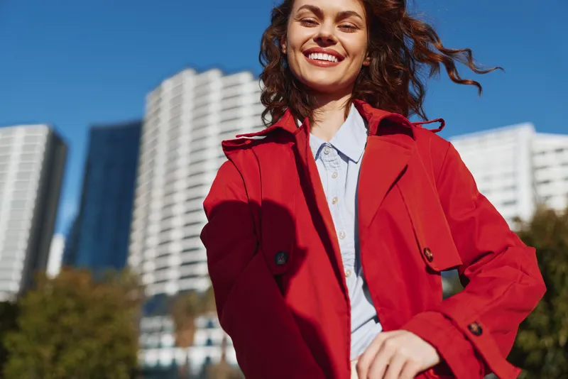 Young woman with curly hair smiles brightly in a red coat outdoors, posing confidently with an urban skyline behind her under a clear blue sky, vibrant colors framing the scene