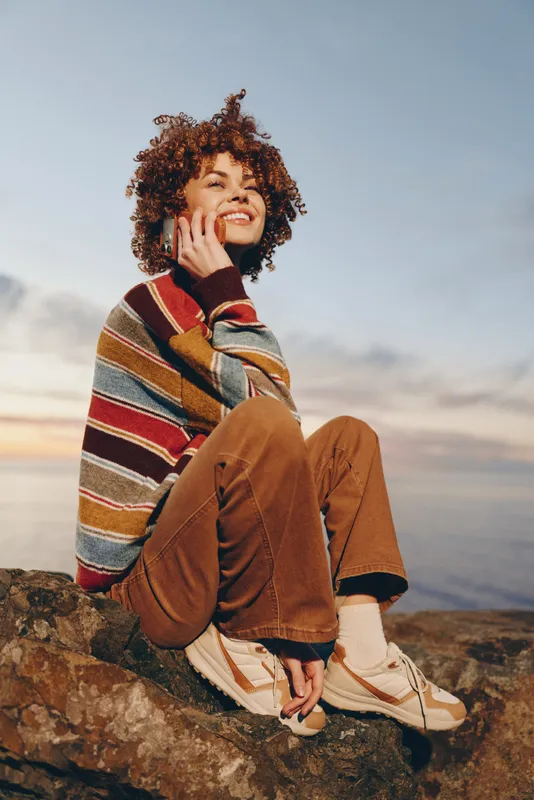 Happy woman outdoor lifestyle on rocky mountains, curly hair blowing, warm sunset landscape sky behind, smile on face, laughing softly, casual jacket and boots, relaxed travel mood