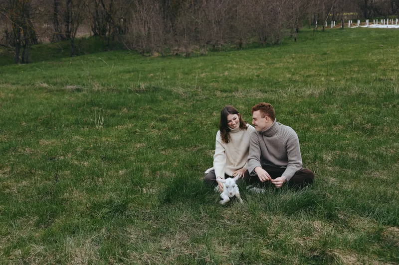 Cheerful couple outdoors on a green meadow, sitting on grass with a small dog, enjoying a relaxed portrait moment, natural light, candid affection, warm breeze, joyful connection and closeness