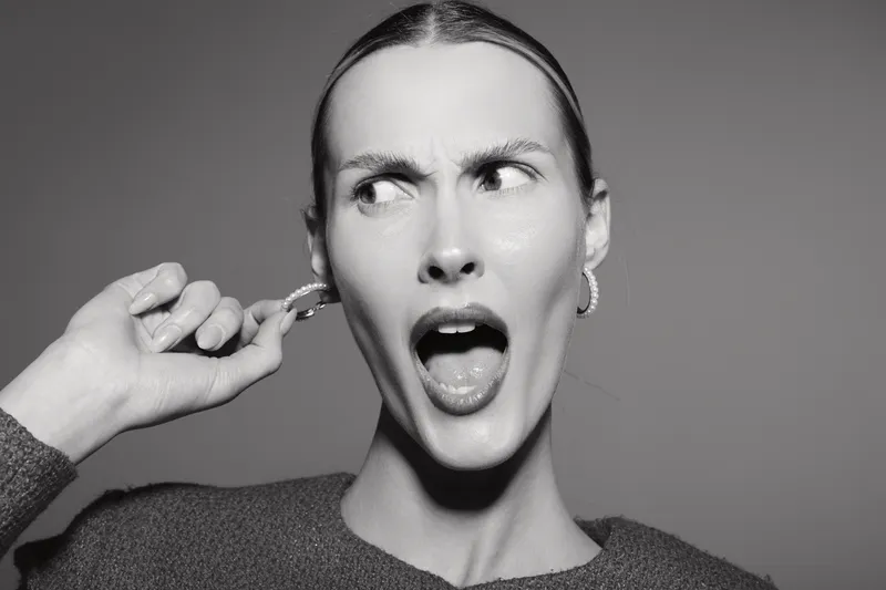 Young white woman with light skin and long hair shows annoyed expression holding finger in ear. Black and white portrait with neutral background. Concept of irritation and discomfort.