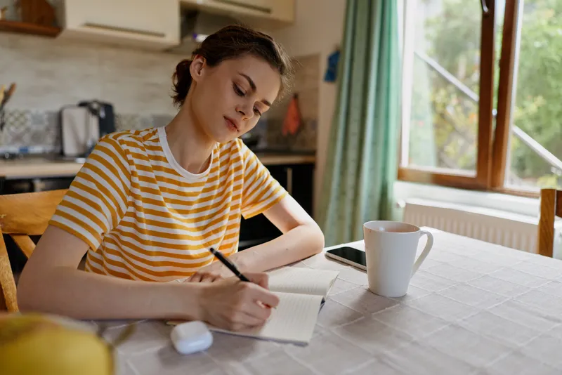 Young woman in a striped yellow t shirt sits at a kitchen table writing in a notebook, creating a calm home moment