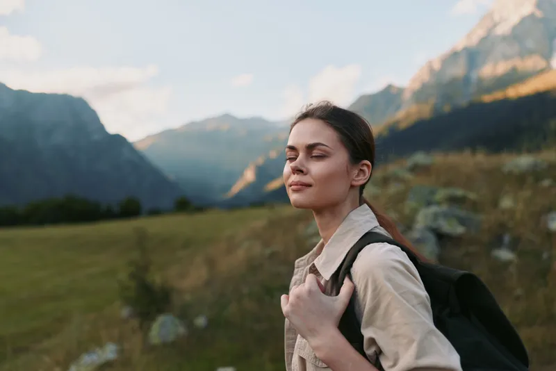 Young woman with a backpack stands in a mountainous landscape with eyes closed and a serene expression, basking in golden light as valley stretches behind her, solitude and exploration mirror her