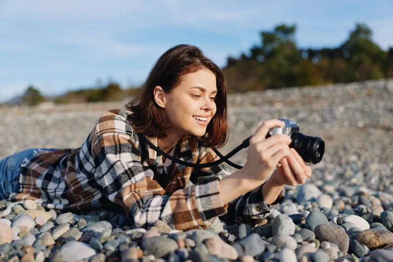 Photographer woman lying on a rocky beach, plaid shirt, capturing a moment with camera under a clear sky, focused and creative in an outdoor scene.