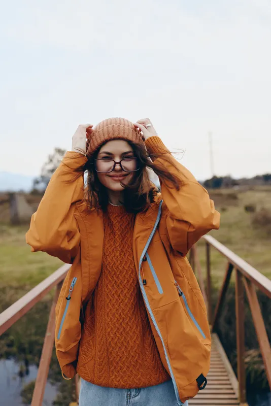 Woman lifestyle outdoors in autumn, orange coat, knitted hat, glasses, sweater, standing on bridge near railing in countryside, hands raised adjusting hat, smiling in windy daylight