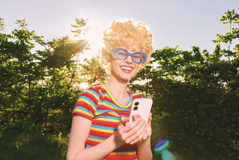 A woman outdoors wearing a colorful striped shirt and blue retro glasses, holding a smartphone, smiling in bright sun, park setting, vintage film color mood