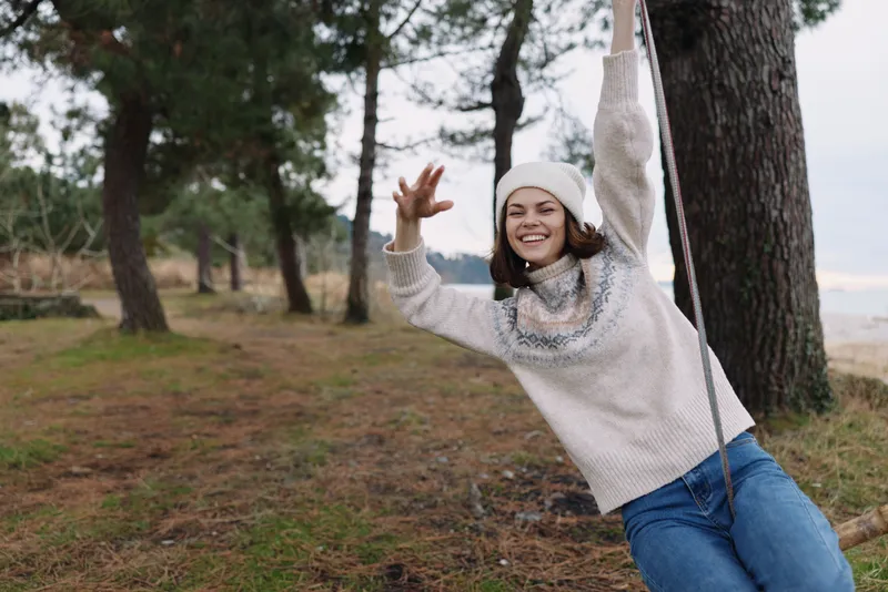 Young woman enjoying a playful moment outdoors, swinging from a rope beside trees in a wooded park, wearing a cozy sweater and knit hat, smiling with a carefree expression and natural light.