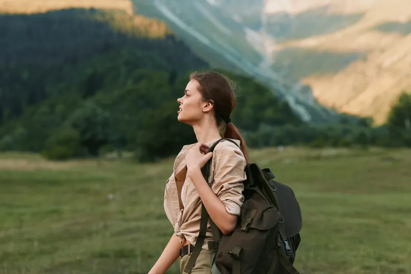 A solo woman with a backpack walks through a grassy field toward distant rugged mountains, conveying a sense of adventure and calm in a wide natural landscape