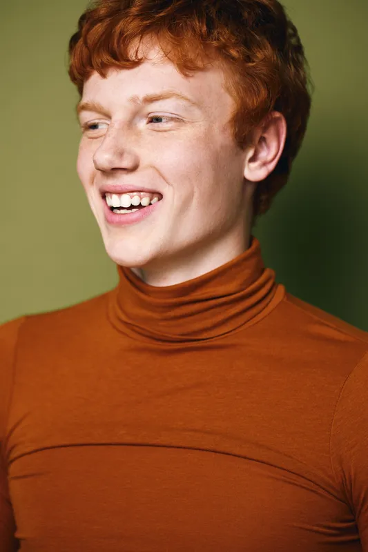 Young man with red hair laughing and looking sideways wearing orange turtleneck sweater against green background, casual portrait with natural expression and curly hairstyle.