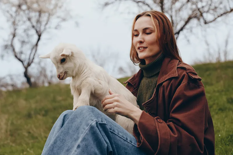 Calm woman in casual attire sits on grass, softly holding a young goat. Outdoor lifestyle scene highlighting care, companionship, and nature under soft daylight.