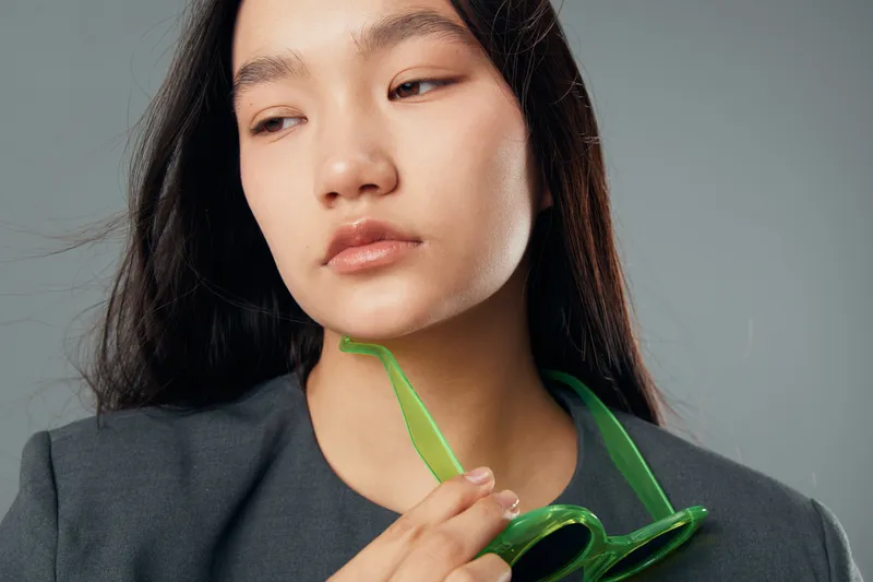 Woman with long dark hair removing green sunglasses looking down thoughtfully against a neutral background in casual outfit for fashion portrait or lifestyle concept.