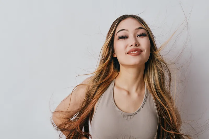 Young woman with long hair and natural makeup posing in casual tank top smiling with confidence against plain background in studio lighting for portrait photography.