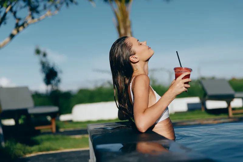 Young woman in white bikini relaxing in outdoor pool holding a red drink with a straw. Sunny day with clear blue sky and blurred garden background. Concept of lifestyle and leisure.