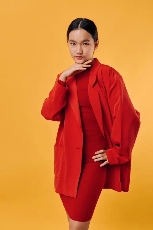 Young woman posing in red dress and oversized blazer on yellow background with serious expression looking at camera for fashion, beauty and style concept portrait.