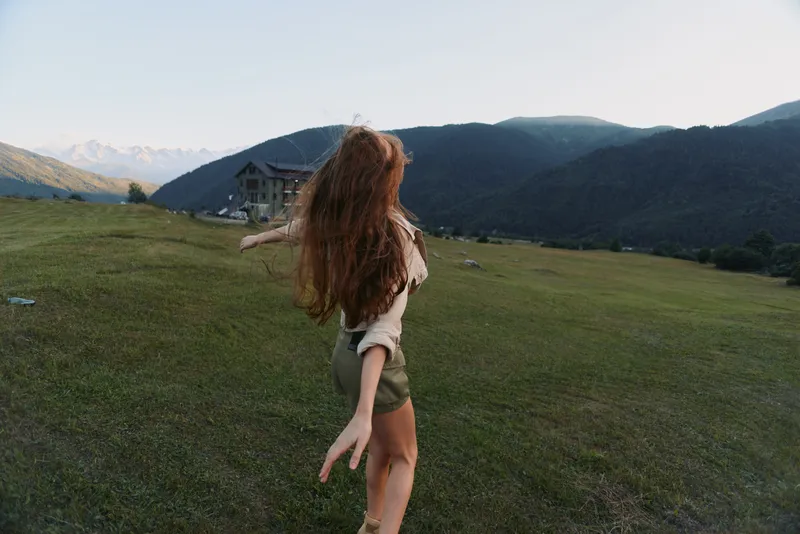 A woman freely explores a grassy hillside, arms outstretched, hair flowing in the breeze. A distant house and mountain range frame a carefree outdoor lifestyle.