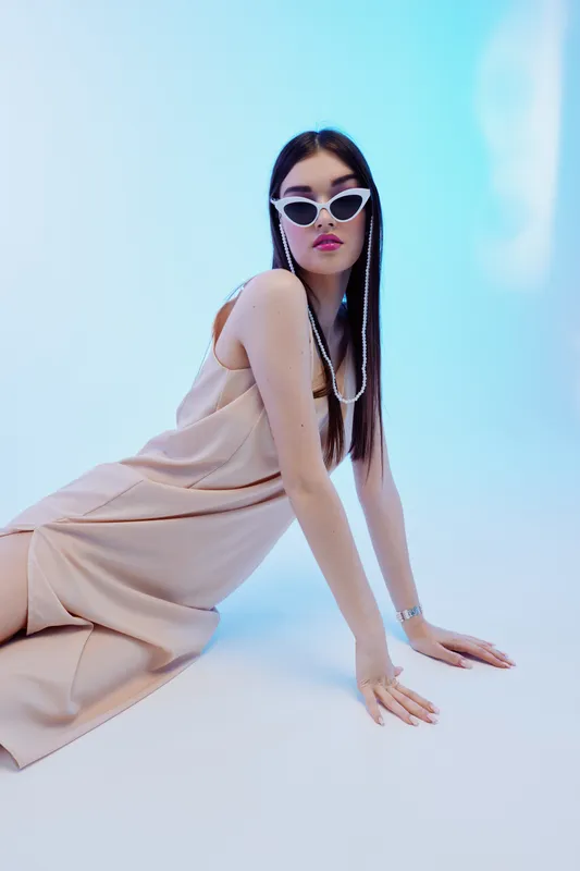 Young woman beauty fashion model wearing beige dress and white sunglasses poses on floor against blue background. Concept for style, elegance, and modern trends in fashion photography.
