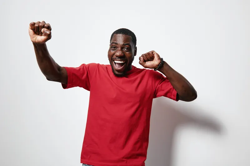 African American man with dark skin wearing red t shirt celebrating with raised fists and joyful expression isolated on white background. Happiness, excitement and success concept.