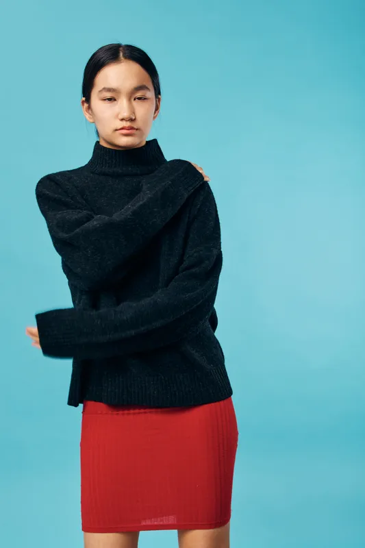 Young woman with long dark hair wearing black knitted sweater and red skirt standing against blue background in studio, casual fashion portrait with natural expression and minimalist style.