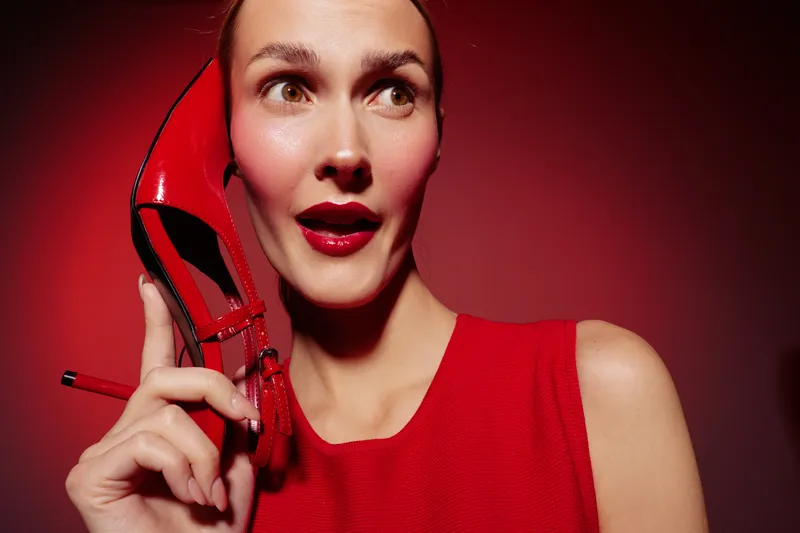 Surprised woman in red dress holds red high heel shoe near face on red background. Concept of fashion, style, glamour, and beauty with vibrant color contrast and studio lighting.