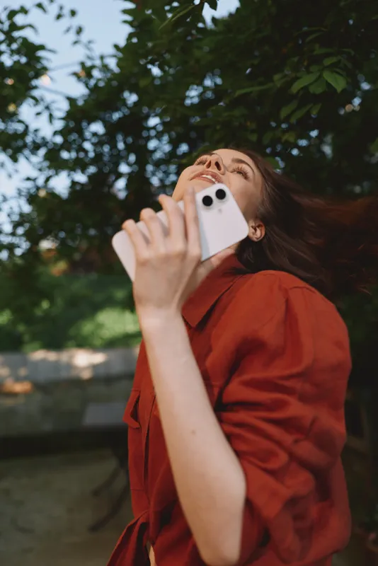 Cheerful woman outdoors, looking up, taking a selfie with a smartphone, sunlit moment, rust red shirt, relaxed pose, lush greenery background