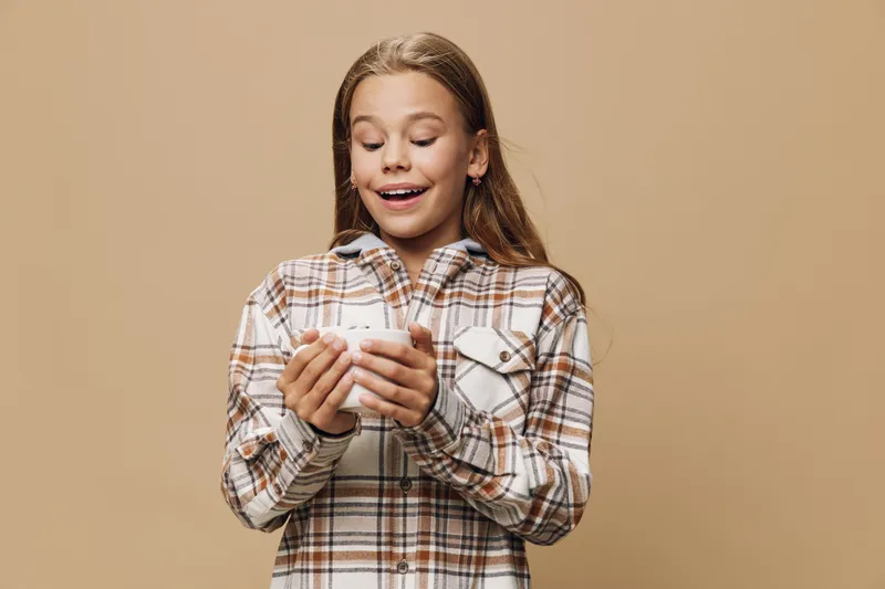 Girl with long hair holding smartphone with both hands looking surprised standing against beige background. Kid wearing checkered shirt using mobile phone for communication or entertainment.