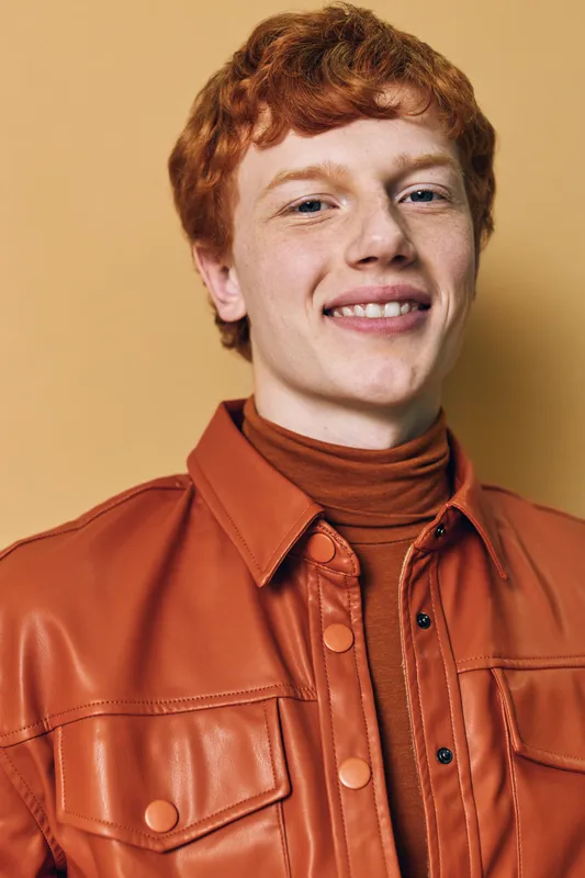 Young man with red hair and freckles smiling, wearing a brown leather jacket and turtleneck sweater against plain background, confident portrait with warm tones.