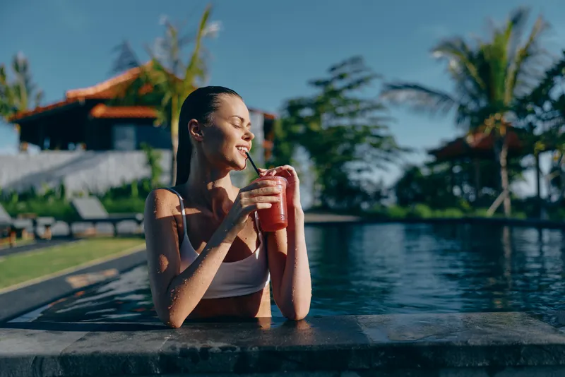 Smiling woman in a white swimsuit lounges at the pool edge, holding a red smoothie glass. Tropical resort backdrop, sunny weather, and relaxed mood convey wellness and leisure.