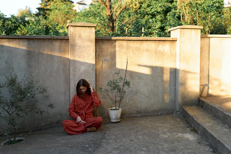 Young woman in a red jumpsuit sits alone on a weathered rooftop terrace beside a potted plant, reflecting in quiet solitude