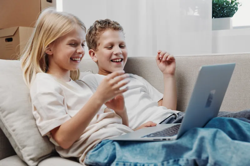Happy children sitting on a sofa using a laptop together, smiling and enjoying digital entertainment in a cozy living room environment. Concept for family, technology, and leisure time.