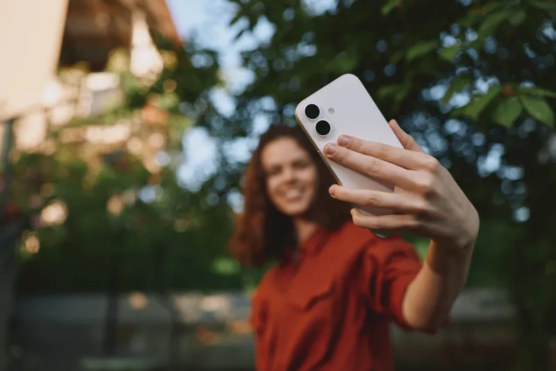 Smiling woman with wavy hair taking a selfie outdoors, casual lifestyle moment, vibrant greenery background, carefree and playful mood, daylight portrait