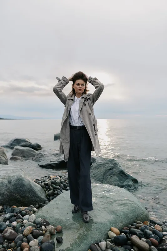Young woman in stylish coat and wide pants stands on rocky shore with hands in hair. Calm sea and cloudy sky create moody atmosphere. Concept of fashion and nature balance.