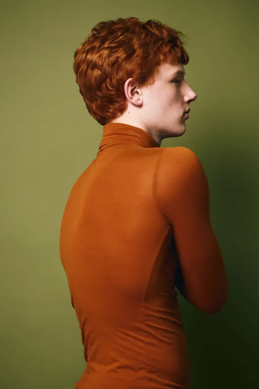 Portrait of young woman with short red hair wearing orange turtleneck sweater standing in profile against green background. Minimal studio fashion photo.