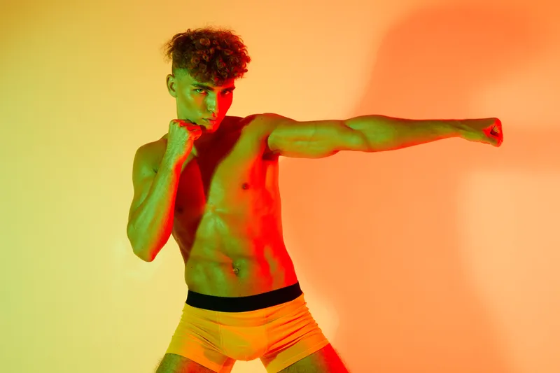 Athletic young man with curly hair practicing boxing stance shirtless in colorful studio lighting for fitness training and martial arts workout.