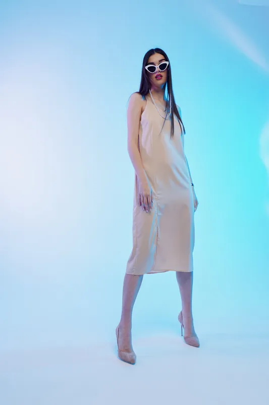 Tall woman in a beige dress and white sunglasses poses confidently in a studio with blue lighting. Concept of beauty, fashion, elegance, and modern style.