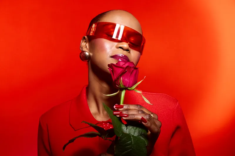 Bald African American woman with dark skin wearing red sunglasses and jacket holds red rose close to face. Studio portrait on red background. Concept of fashion, beauty, and self-expression.