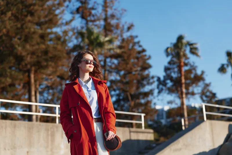 A fashionable woman wearing a red coat and sunglasses stands outdoors on a sunny day, posing with hands in pockets and exuding confident, stylish charm.