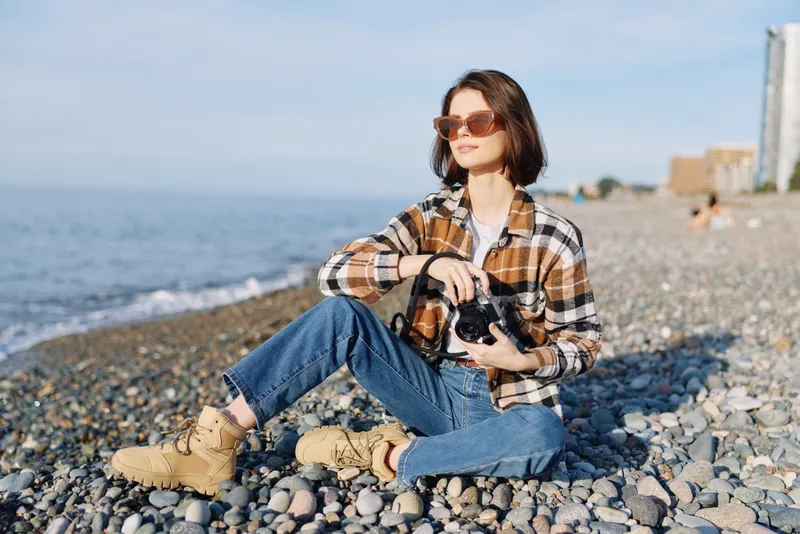Photographer woman sitting on a pebble beach with a camera, wearing a plaid shirt, jeans and sunglasses, relaxed outdoor scene capturing coastline moments and casual seaside photography