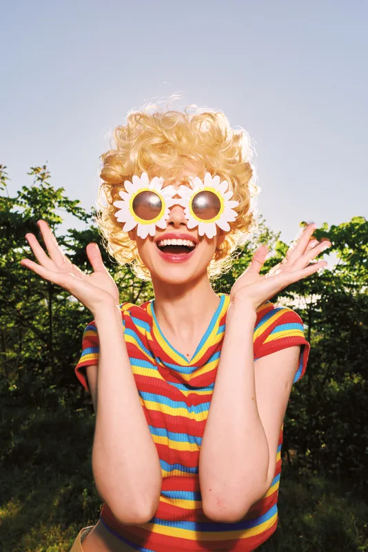 Playful woman outdoors wearing a striped shirt and sunflower goggles, arms raised in a joyful pose. Retro film color and bright light create a warm, nostalgic mood and carefree lifestyle.