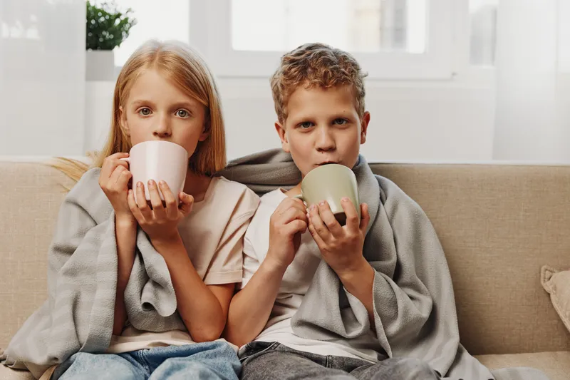 Two children wrapped in blankets sit on a couch drinking hot beverages. Cozy indoor scene with natural light, capturing comfort, warmth, family bonding, and relaxation during cold weather.
