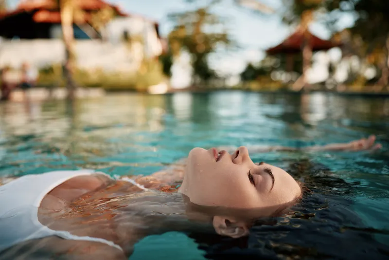 A woman floats on her back in a sunlit pool, eyes closed, enjoying tranquil water and warm breeze. A lifestyle moment of relaxation, selfcare, and peaceful summer escape.