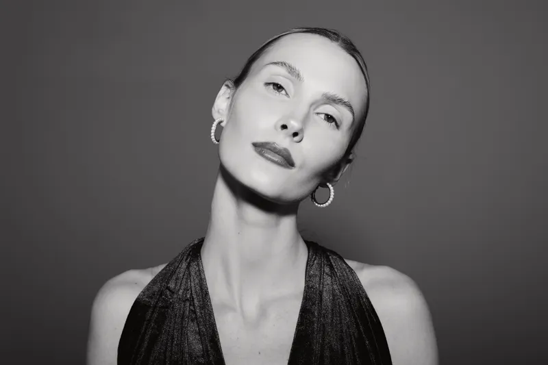 Elegant Caucasian woman with pale skin and slicked back hair poses confidently in a shiny black dress and hoop earrings on grey background. Fashion and beauty concept.