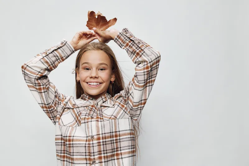 Girl smiling and holding autumn leaf above head in plaid shirt on neutral background. Young happy child with long hair posing for portrait in casual clothes with fall foliage.