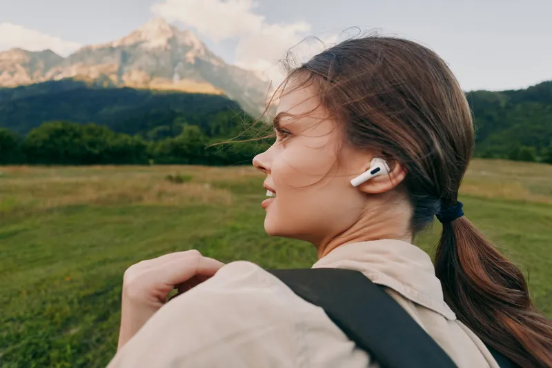 Young woman with ponytail wearing wireless earbuds hiking in green meadow with mountain background enjoying nature and active lifestyle outdoors in fresh air.