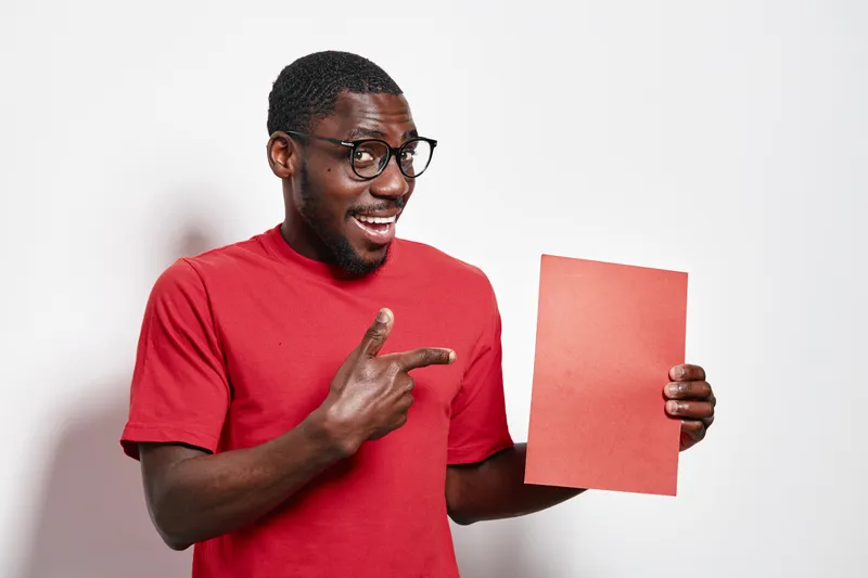 African American man with dark skin wearing glasses and red shirt pointing at blank red paper for presentation or advertisement on white background. Smiling, casual and confident.