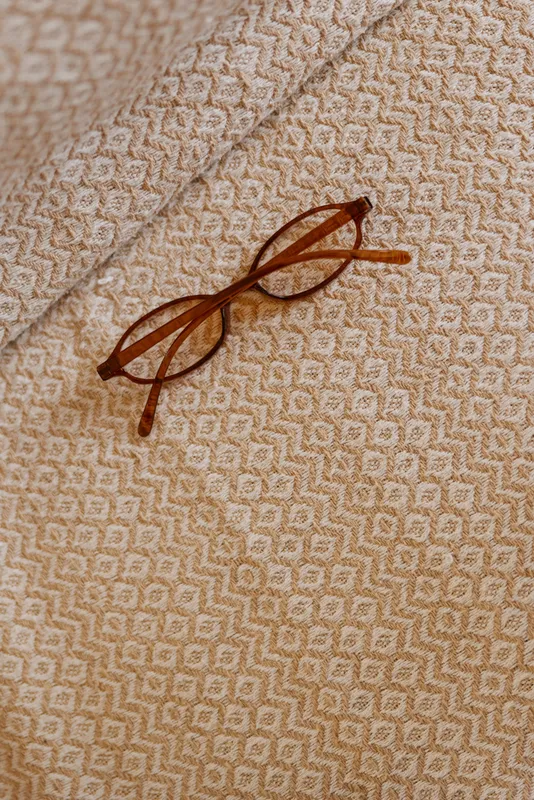 A pair of brown glasses rests on a beige textured fabric with a subtle diamond pattern, evoking calm, focus, and intimate home study moments in a cozy setting.