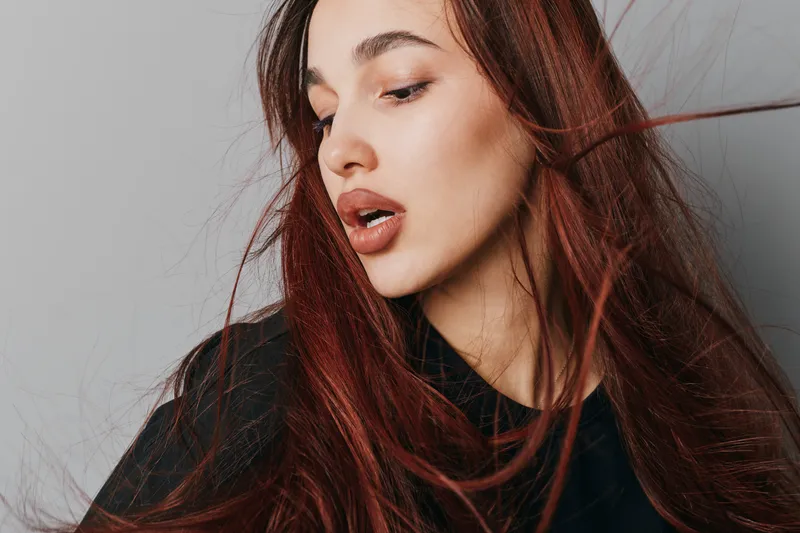 Woman with long red hair and closed eyes posing in studio with neutral gray background. Female model with natural makeup and flowing hair wearing black top for beauty portrait.