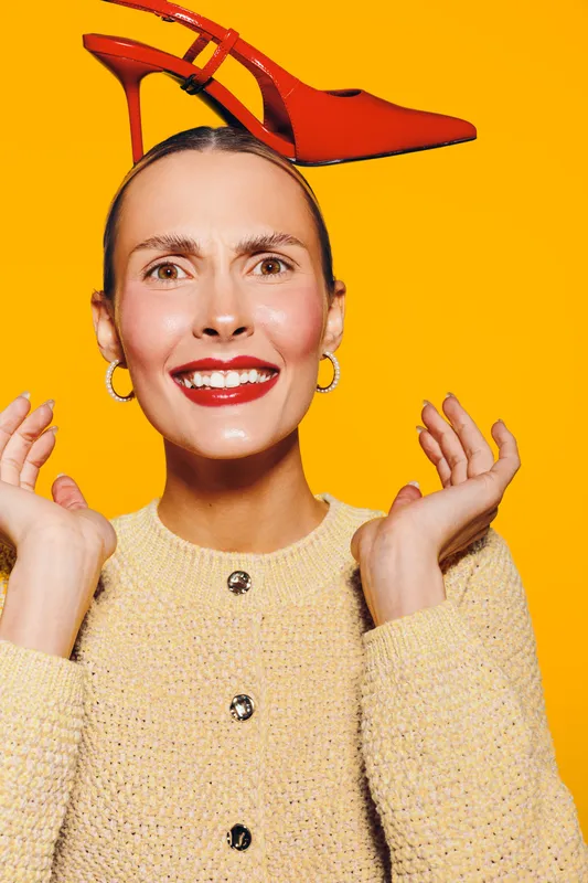 Smiling woman with red high heel shoe balanced on her head wears beige sweater and gold hoop earrings. Bright yellow background creates playful and quirky fashion concept with confident mood.