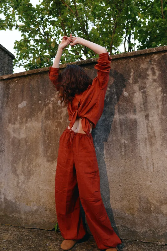 Woman outdoor red clothing casual lifestyle pose fashion street wall back to camera with raised arms sunlit scene and leafy backdrop