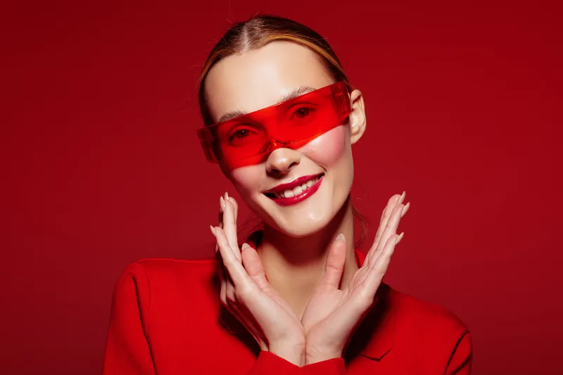 Smiling woman in red sweater and red sunglasses poses against red background. Bright studio lighting highlights her joyful expression and stylish fashion concept for modern lifestyle.