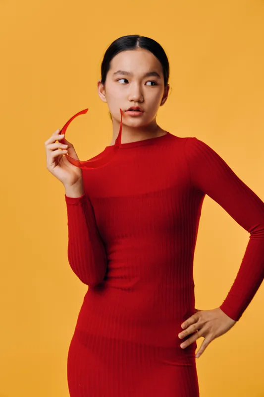Young woman in red dress holding red glasses near face looking to side posing on orange background. Stylish model with black hair tied back wearing long sleeve outfit for fashion portrait.