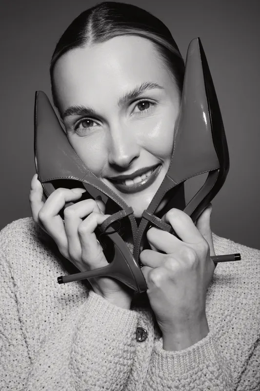 Smiling white woman with light skin holding red high heel shoes close to her face in black and white studio portrait. Concept of fashion, beauty, and happiness.